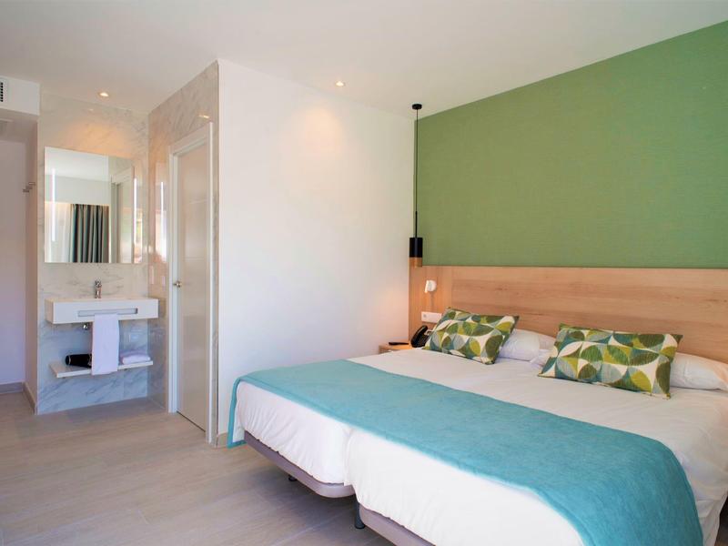 Habitación de hotel moderna con cama doble, acento verde en la pared y área de baño abierta.