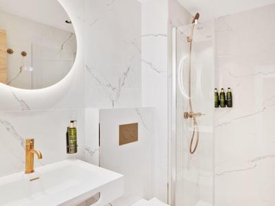 Baño moderno con espejo redondo iluminado, cerámica blanca y grifería dorada.