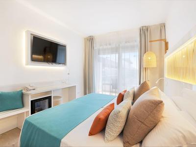 Habitación de hotel luminosa con cama doble, cojines azules y naranjas, televisor de pared y ventana grande.