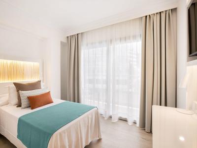 Habitación de hotel luminosa con cama individual, cortinas beige y ventana grande con luz natural.