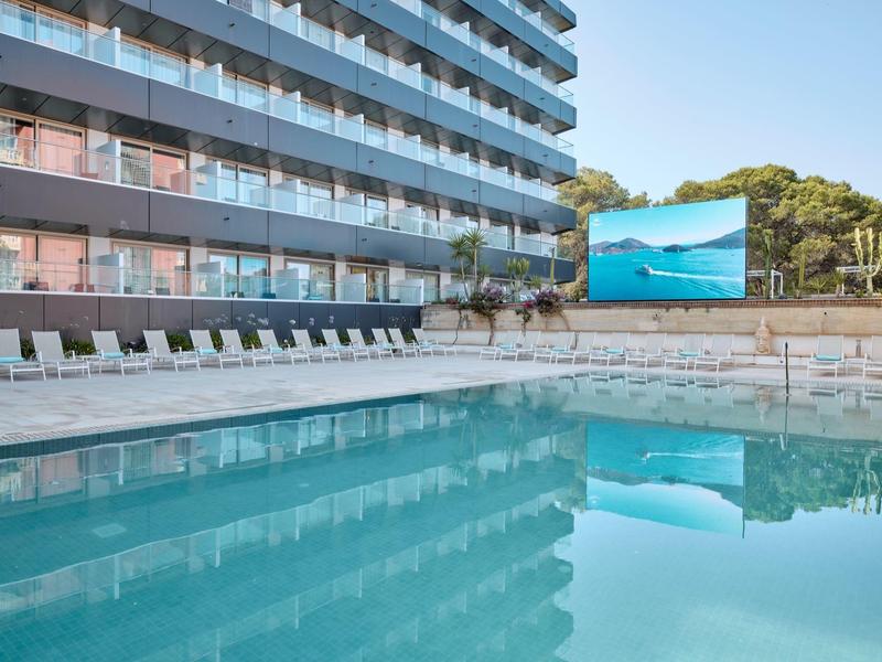 Piscina al aire libre con tumbonas junto a un edificio moderno de hotel de varios pisos.