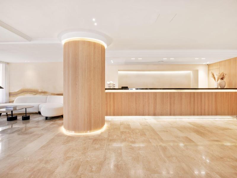Área luminosa del vestíbulo del hotel con mostrador largo de recepción de madera y una columna redonda de madera en el centro.