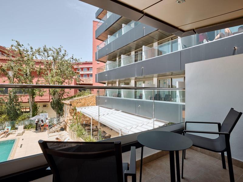 Balcón con mesa y sillas negras, vista a piscina y edificio de hotel con balcones de vidrio.