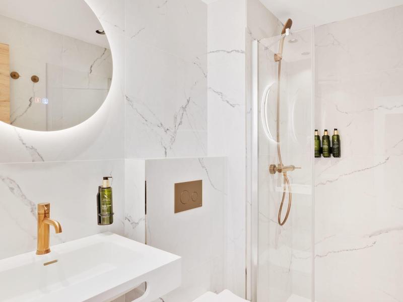 Baño moderno con espejo redondo iluminado, cerámica blanca y grifería dorada.