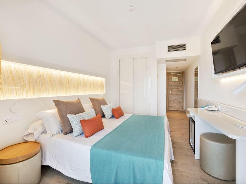 Habitación de hotel moderna con cama doble, cubrecama azul y TV en la pared en ambiente claro.