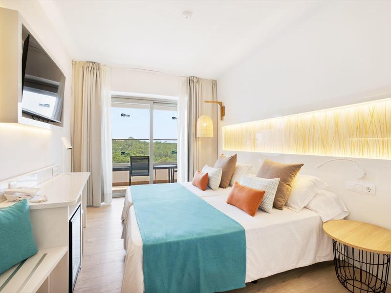 Habitación de hotel luminosa con cama doble, acentos azules y balcón con vista a la naturaleza.