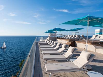 Luxusní lehátka s modrými slunečníky u moře s plachetnicí v pozadí.