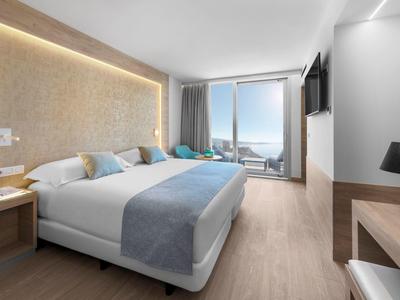 Moderní hotelový pokoj s velkou postelí, dřevěnou podlahou a výhledem na moře přes velká okna.