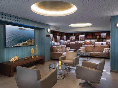 Moderní hotelový lounge s posezením, velkou obrazovkou a centrálními stoly za tlumeného osvětlení.