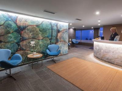 Moderní hotelová lobby s dekorativní kamennou skleněnou stěnou, modrými křesly a světlou recepcí.