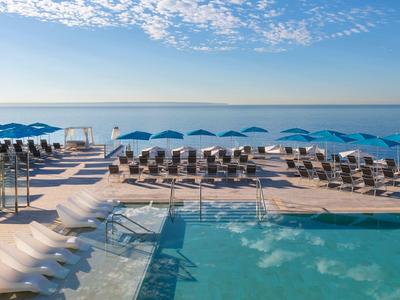 Moderní bazénová část s lehátky a modrými slunečníky s výhledem na moře pod modrou oblohou.