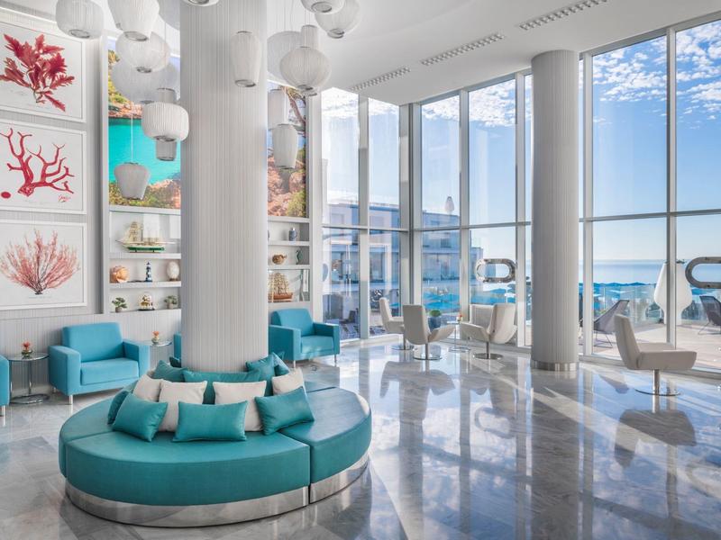 Světlý hotelový lounge s kruhovým posezením a výhledem na moře skrz velká okna.