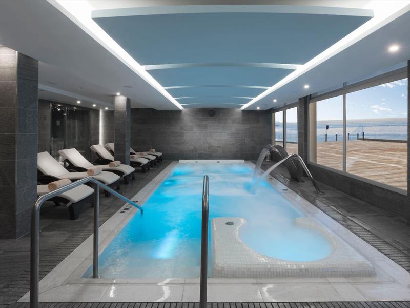 Moderní vnitřní bazén s vířivkou a lehátky, velká okna s výhledem na pláž a moře.