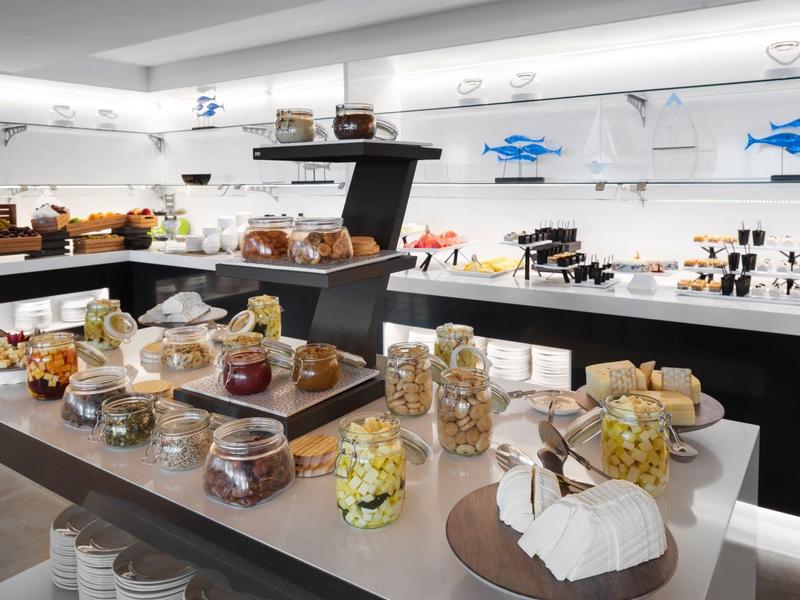 Moderní snídaňový bufet se sýry, chlebem a pomazánkami v světlé místnosti.