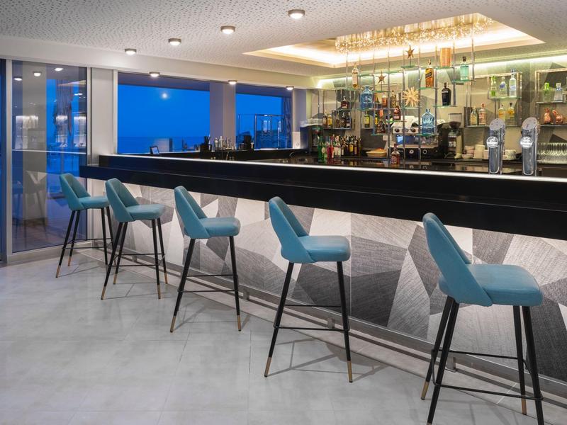 Moderní hotelový bar s modrými barovými židlemi a geometrickým designem pod teplým osvětlením.