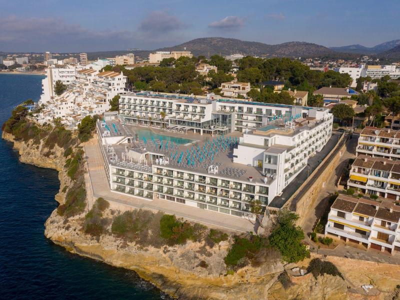 Velký hotel s několika bazény na útesu u moře s okolními budovami a kopci.