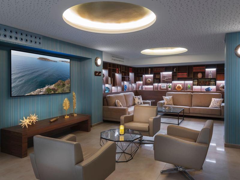 Moderní hotelový lounge s posezením, velkou obrazovkou a centrálními stoly za tlumeného osvětlení.