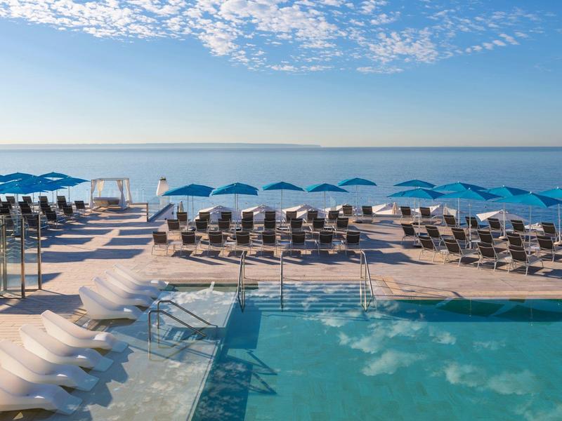 Moderní bazénová část s lehátky a modrými slunečníky s výhledem na moře pod modrou oblohou.