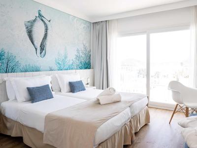 Helles Hotelzimmer mit zwei Betten, Meerbild an der Wand und großem Fenster mit Vorhängen.