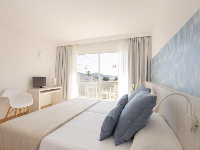 Jasny pokój hotelowy z podwójnym łóżkiem, szarymi poduszkami, telewizorem i balkonem z widokiem.