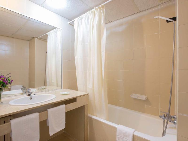 Jasna łazienka hotelowa z umywalką, lustrem, kwiatami i wanną z zasłoną prysznicową.