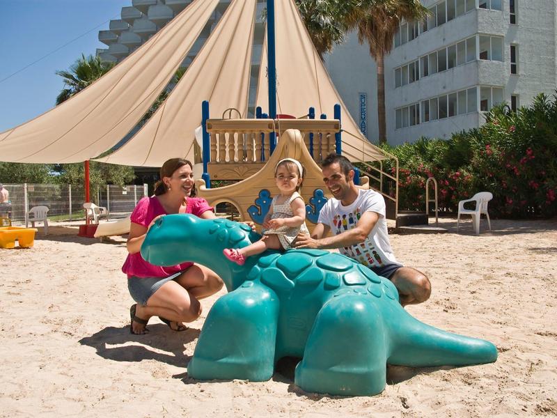 Troje dorosłych bawi się dużą zieloną strukturą do zabawy w kształcie krokodyla na piaszczystej plaży przed hotelem.