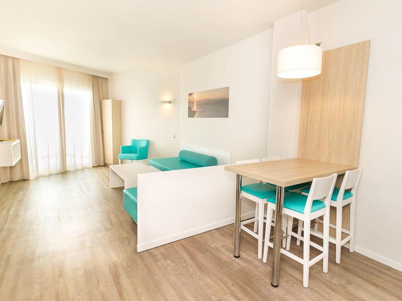 Jasny, nowoczesny salon z drewnianą podłogą, turkusowymi meblami i dużymi oknami z zasłonami.