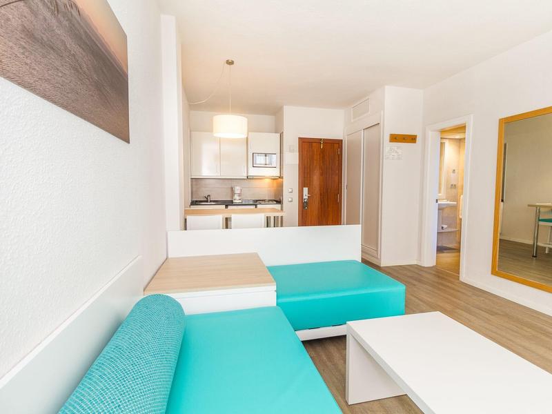 Nowocześnie umeblowany pokój hotelowy z turkusową sofą i aneksem kuchennym.