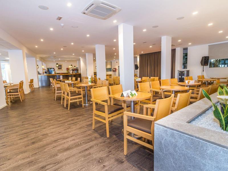 Jasna jadalnia hotelowa z drewnianymi podłogami, drewnianymi stołami i krzesłami, nowoczesnym oświetleniem i zielonymi roślinami.