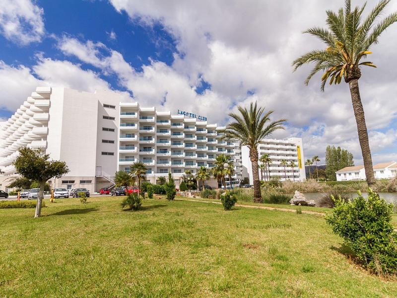 Duży budynek hotelu z balkonami, otoczony zieloną trawą i palmami pod niebieskim niebem.