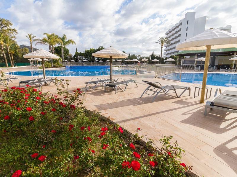 Basen hotelowy z leżakami, parasolami i kwiatami na pierwszym planie pod pochmurnym niebem.