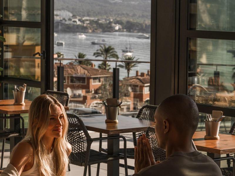Due persone sedute in un caffè con vista su una città costiera e montagne sullo sfondo.