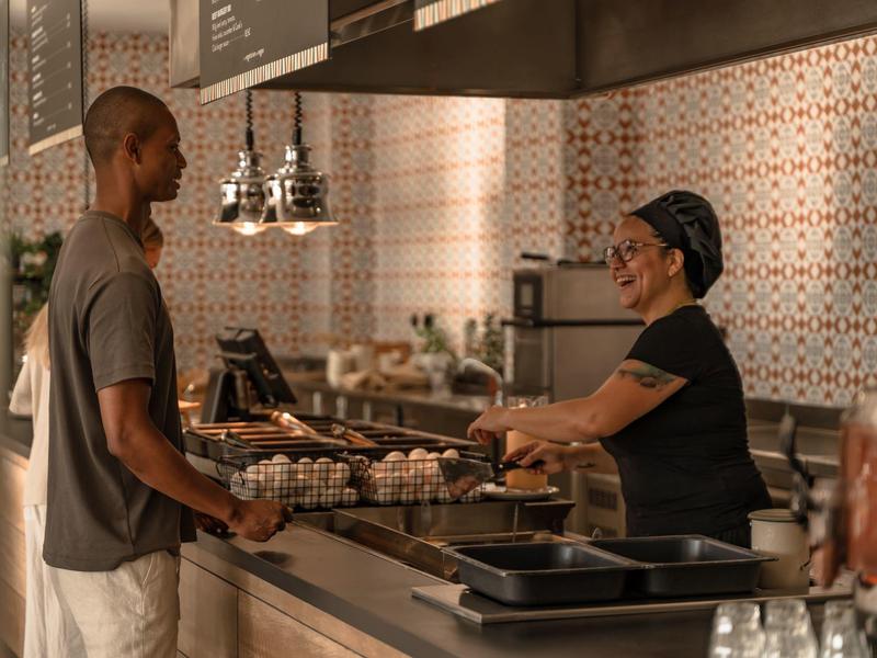 Persona che ordina in un bar con piastrelle decorate sul muro e una cassiera dietro il bancone.