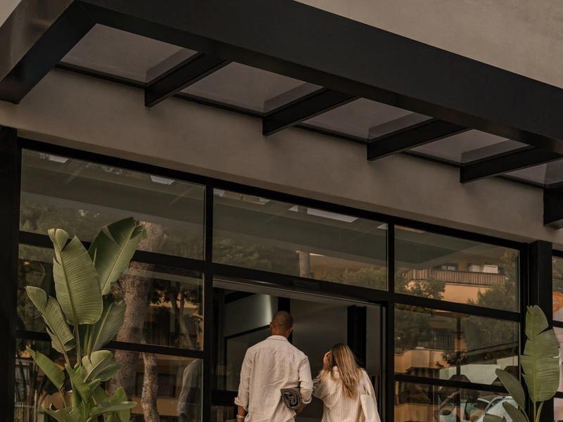 Due persone con abiti eleganti entrano in un caffè con grandi vetrate e piante.