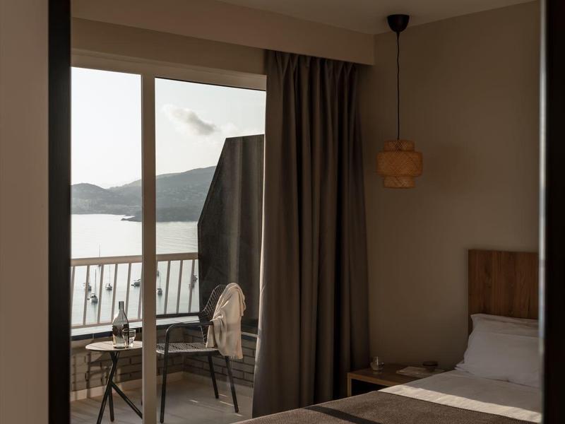 Camera d'hotel semplice con letto, tende e balcone con vista su acqua e colline.