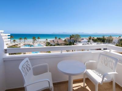 Balcon avec petite table blanche et deux chaises, vue sur piscine, palmiers et mer sous un ciel clair.