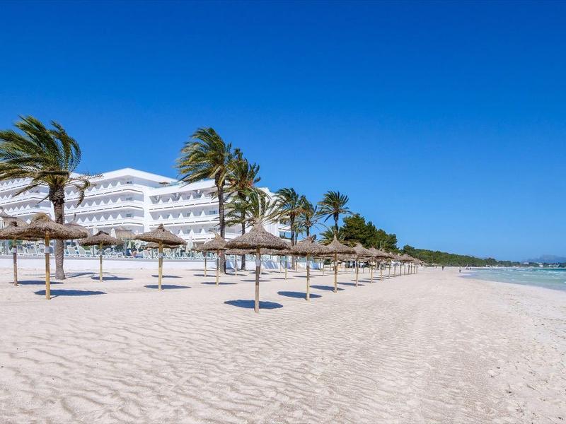Large plage de sable avec des palmiers et des parasols devant un grand hôtel blanc au bord de la mer.