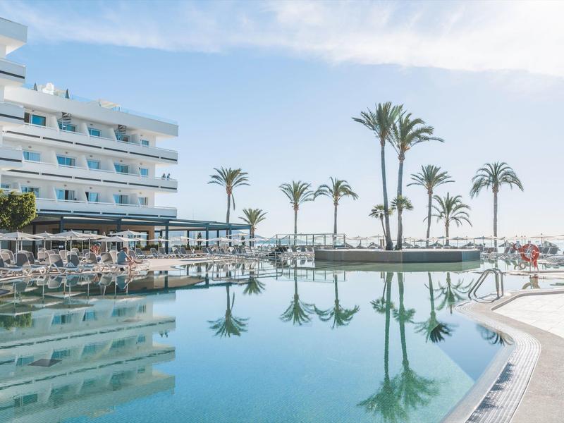 Grand piscine d'hôtel avec des palmiers et des transats sous un ciel bleu.