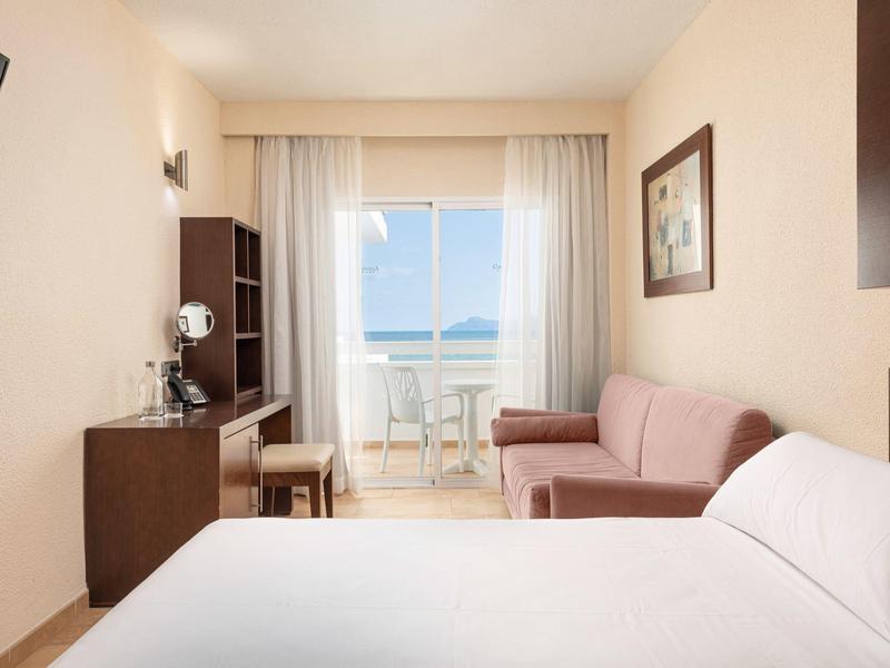 Chambre d'hôtel lumineuse avec un lit double, un canapé, un bureau et un balcon avec vue sur la mer.