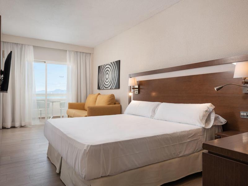 Chambre d'hôtel lumineuse avec grand lit, coin salon et balcon avec vue sur la mer.