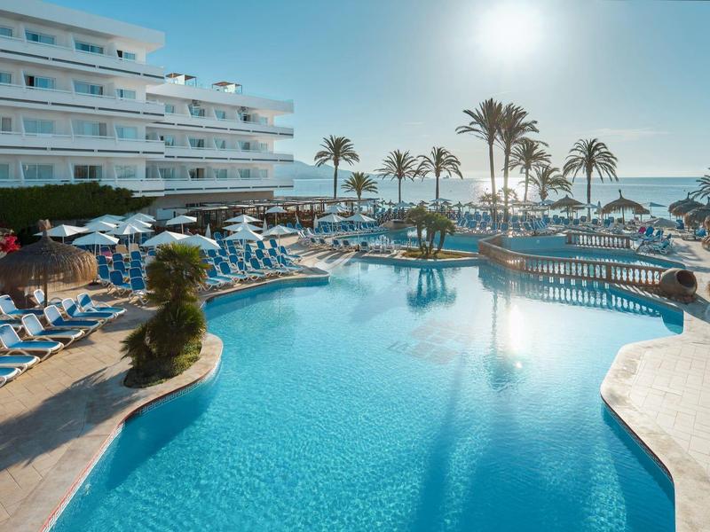 Grand piscine d'hôtel avec transats et palmiers sous un ciel ensoleillé au bord de la mer.