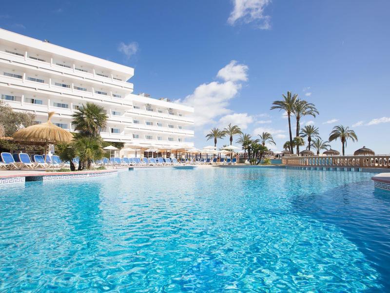 Grande piscine d'hôtel avec chaises longues et palmiers sous un ciel bleu clair.