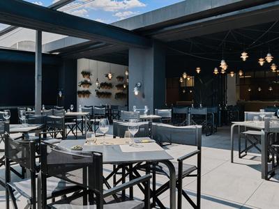 Moderner Außenbereich eines Restaurants mit schwarzen Stühlen und Tischen, heller Boden und blauer Himmel.