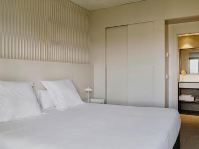 Schlichtes Schlafzimmer mit Bett, weißen Kissen, minimalistischer Wandgestaltung und Blick ins Badezimmer.