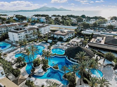 Aerial view of a hotel with multiple pools, palm trees, and a view of the sea and mountains in the background.