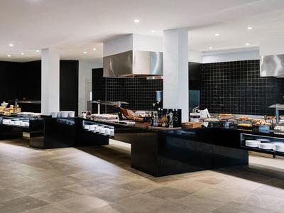 Moderne Großküche mit schwarzen Arbeitsflächen, Edelstahlgeräten und weißen Säulen unter Deckenbeleuchtung.