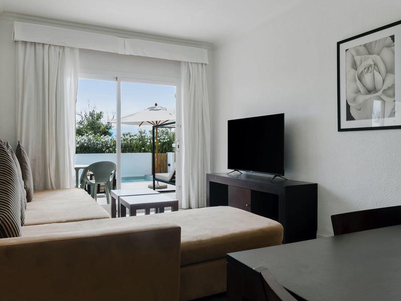Helles Wohnzimmer mit braunem Sofa, TV auf schwarzem Sideboard, großem Fenster und Balkon mit Meerblick.