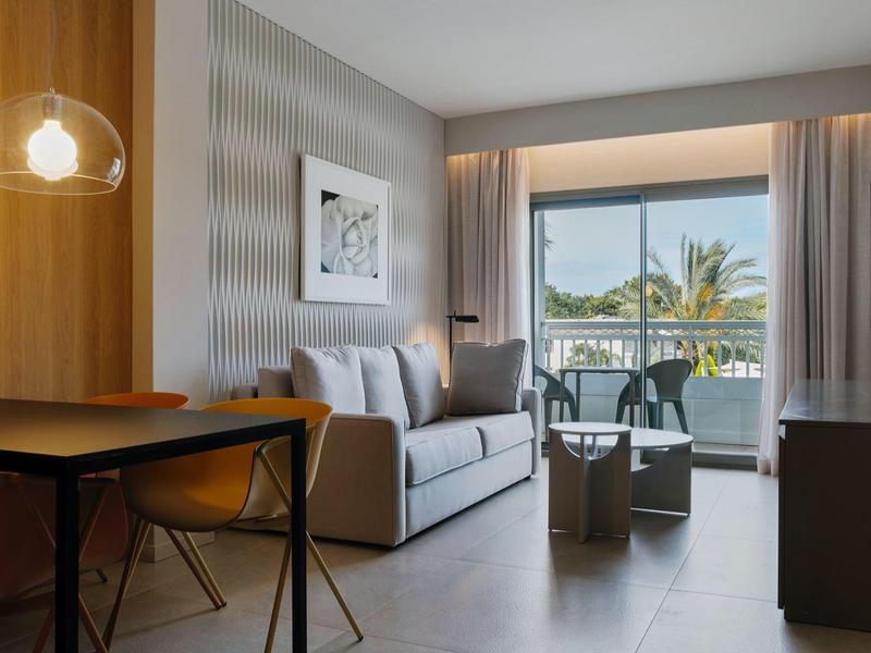 Modern eingerichtetes Wohnzimmer mit grauem Sofa, Holztisch, Glastür und Balkon mit Palmenblick.