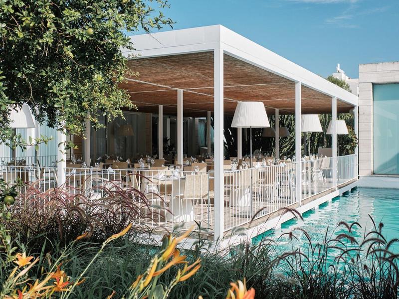 Modernes, offenes Restaurant mit weißen Stühlen, großem Dach und Pool neben grünen Pflanzen.