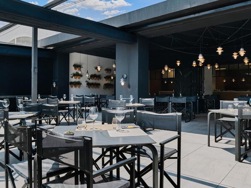 Moderner Außenbereich eines Restaurants mit schwarzen Stühlen und Tischen, heller Boden und blauer Himmel.